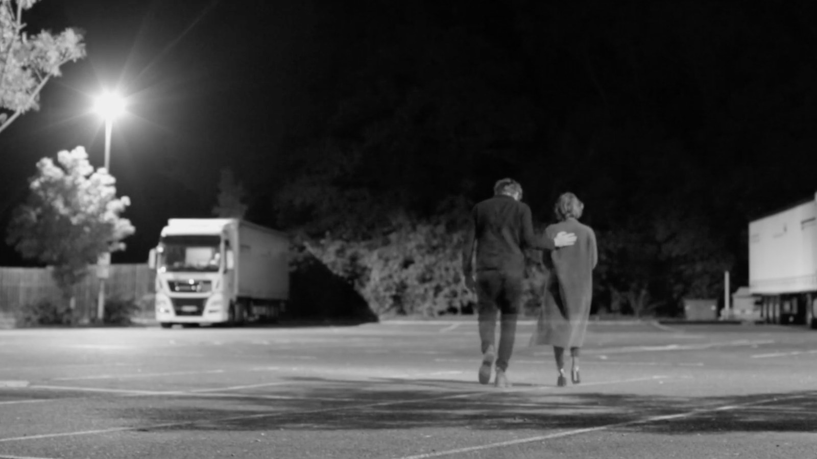 Two figures crossing a lorry park at night, black and white